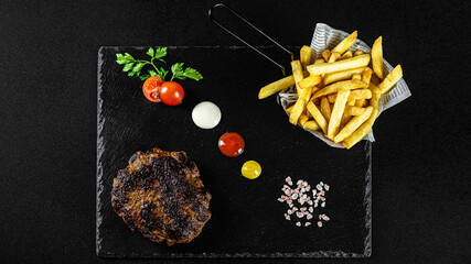 Grilled spicy Gurmanska Pleskavica  with cheese, fries in a basket on a black plate with sauces and fresh veggies