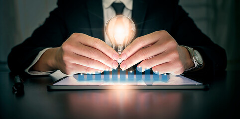 businessman hand holding bright light bulb with graph economic growth success chart on tablet. Business hand showing creative business strategy with light bulb and graph as concept.