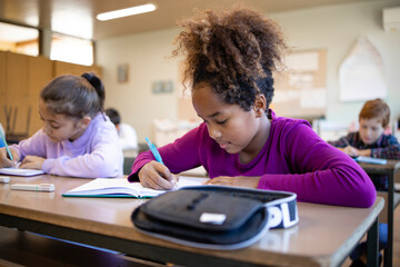 Concentrated schoolgirl sitting at desk and writing in exercise book with classmate sitting next to...