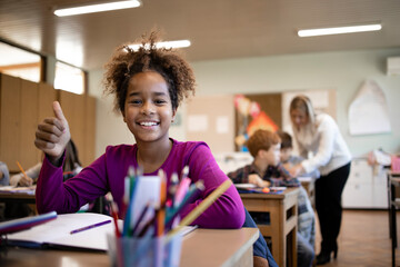 Portrait of smiling Afro American school child sitting in classroom with classmates and teacher in background. - obrazy, fototapety, plakaty