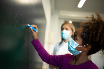 School girl wearing face mask and writing on board under teacher's supervision during corona virus measures.