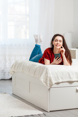 A young woman is lying on the bed on her stomach, holding a phone and smiling. White interior of the room in the background.Copy space. Vertical. Concept of communication and social networks