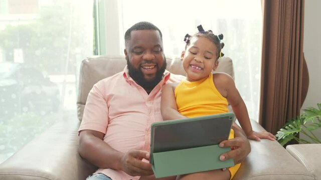 Happy family African father and little daughter sit on sofa using digital tablet playing game or watching movie together. Dad and child girl kid enjoy leisure activity with wireless technology at home