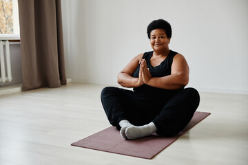 Fototapeta premium Minimal full length portrait of active senior woman sitting in lotus position while enjoying yoga indoors and smiling at camera