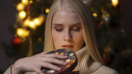 Young happy woman opening present box close-up. Beautiful model holding christmas gift, lying on sofa in decorated living room. Concept of new year holidays, cozy winter evening.