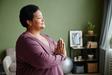 Side view waist up portrait of black senior woman meditating at home with eyes closed - obrazy, fototapety, plakaty