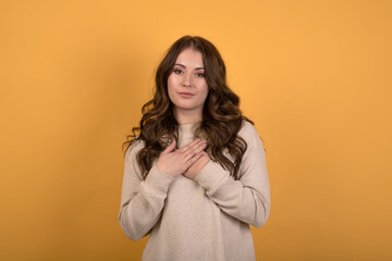 A beautiful and attractive Caucasian brunette girl in a casual jumper showing gratitude while holding her hands on her chest on an orange studio background.