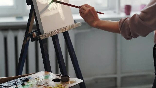 Art school. Female student. Artwork creation. Unrecognizable woman painting on canvas with brush in light room interior.