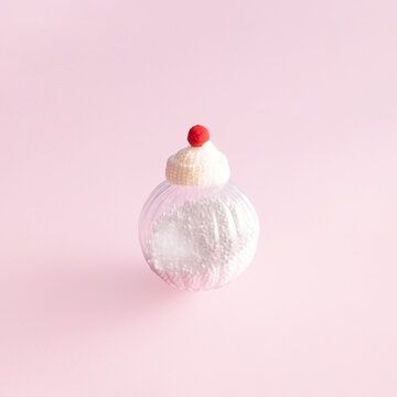 Decorative Christmas bauble, snow inside, woolen hat with pompom on top. Minimal creative holidays layout against pastel pink background.