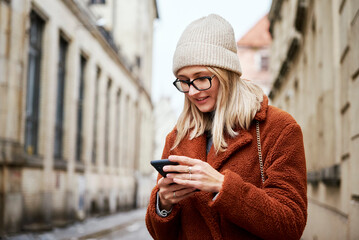 Beautiful woman in coat using smartphone at city street. Online communications