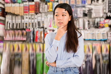 Portrait of cheerful glad Asian girl having doubts about choice of goods in cosmetics store