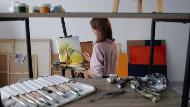 Artwork creation. Female painter. Inspiration muse. Happy woman painting scenery view with trees on canvas in light studio interior.