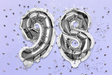 Silver foil balloon number, digit ninety eight on a lilac background with sequins. Birthday greeting card with inscription 98. Top view. Numerical digit. Celebration event, template.