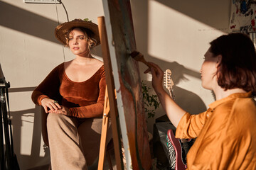 Brunette woman artist drawing with brush on canvas while her girlfriend model sitting