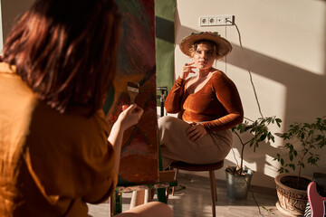 Woman posing to her girlfriend while she sitting in front of the easel and drawing
