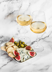 Appetizer plate with blue cheese, olives, homemade cheese cracker, figs  and two glasses of white wine on a light marble background, top view