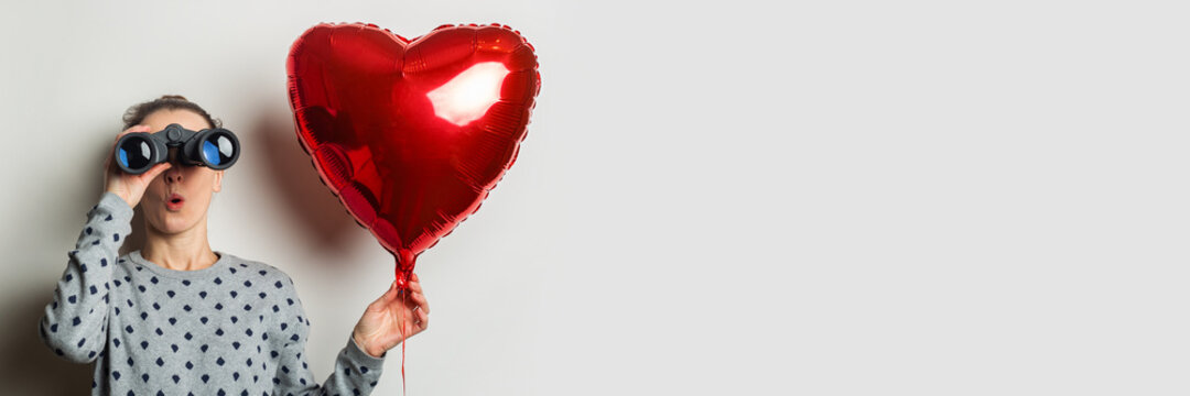 Young woman in a sweater looks through binoculars and holds an air balloon heart on a light background. Valentine's Day concept. Search for a loved one