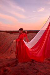 Beautiful young woman or girl with curly hair and in red dress with a light flying fabric on the sand on sunny day with blue sky in the background. Model or dancer posing in photo shoot on sand dunes