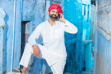 Happy North Indian mature man with beard and red turban talking on smart phone, Smiling Traditional male calling on android smart mobile phone while standing outdoor against blue background,