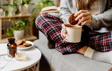 Cozy woman legs in knitted winter warm socks, sweater and checkered pajama drinking hot cocoa or coffee in mug, sitting on couch at home. Autumn mood with candle, decor, cookies and indoor plant