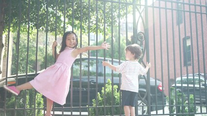 slow motion shot of a little boy and a little girl playfully hanging on the black railing in the courtyard with cars parked behind the railing on a bright sunny day. - Powered by Adobe