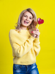 Studio portrait of a beautiful blonde. model in yellow sweater and blue jeans posing for the camera, having fun, balust, laughing with lollipop, candy heart. yellow, colored background