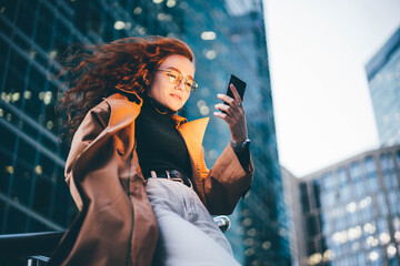 Successful woman using smartphone outdoors while standing near skyscraper at night.