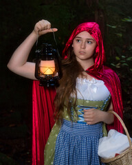 Teenage girl dressed as Little Red Riding Hood holding a lit lantern near her face as she looks out...