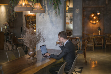 Young caucasian business man in formal suit working on laptop while drinking coffee in a bar. Handsome man working at a cafe, typing on keyboard