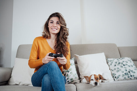 Beautiful woman playing video game sitting on sofa with dog at home
