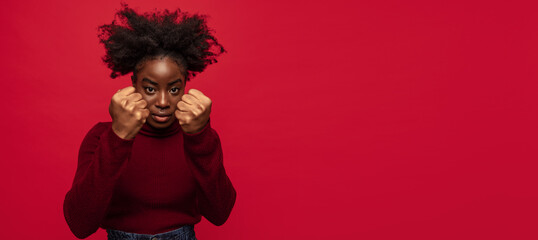 Displeased African young girl showing her fists as symbol of protection from violence isolated on dark red background.