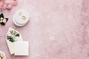 Studio shot of invitation card, ribbon, engagement ring and pair of wedding rings flat laid against pink background