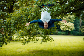Playful businessman with arms outstretched hanging upside down from tree at park