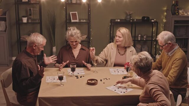 High-angle slowmo shot of five senior men and women sitting around dinner table in evening playing lotto and having conversation