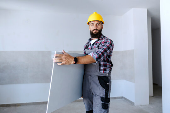 A strong bearded worker in overalls and with a helmet on head relocates plasterboard. It is ready to be put on the ceilings and walls.