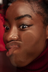 Fototapeta premium Close-up face of funny dark skinned young girl crushed on glass isolated on dark red studio background.