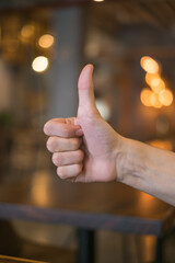 hand showing thumbs up sign against the brown backgrounds with lights in coffee shop. High quality photo