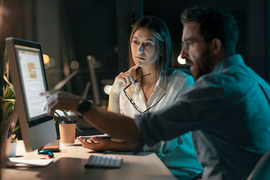 Thoughtful businesswoman by colleague explaining strategy over computer at night in office