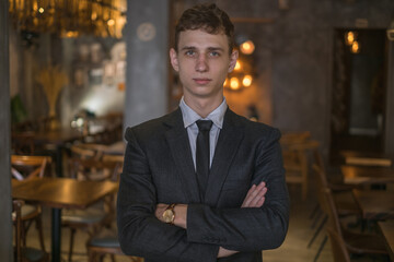 Portrait of young caucasian business man in formal suit standing in the bar with crossed arms. Handsome man standing in the coffee shop 