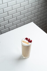 Tall latte in a glass with latte art and red flowers on top, white cafe table