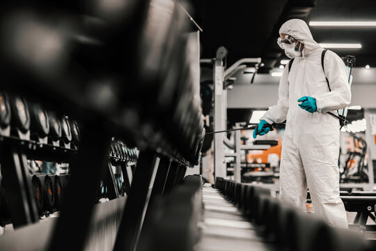 Suppression Of COVID-19 Virus And Cleaning Of Neatly Arranged Exercise Equipment In The Gym By A Male Person In A Protective White Suit. Antiseptic, Disinfection, Cleanliness