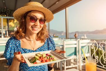 A smiling woman in a hat with a plate of Greek salad on the terrace of a restaurant by the sea....