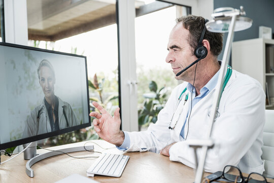 Doctor wearing headset discussing with colleague on video call through computer at desk