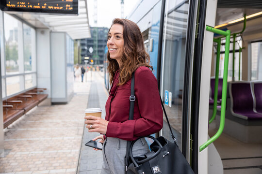 Smiling businesswoman holding disposable cup and mobile phone disembarking from train at station