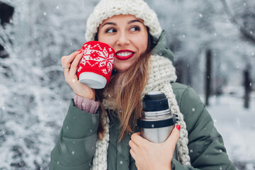 Woman drinking hot tea holding vacuum flask in snowy winter park. Cup dressed in red knitted Christmas case