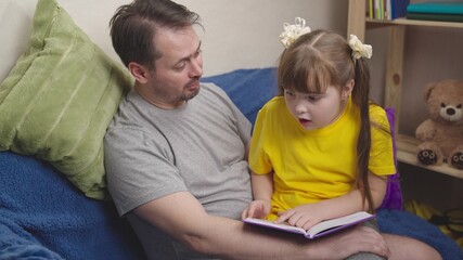 cheerful dad with kid daughter reading a book at home while sitting on couch about embracing each other, child teaches lessons with his father remotely, happy family, getting school education at home