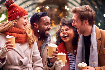 Group Of Friends Drinking Hot Chocolate With Marshmallows In Snow At Outdoor Christmas Market