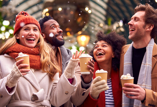 Group Of Friends Drinking Hot Chocolate With Marshmallows In Snow At Outdoor Christmas Market