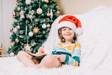 Obraz na płótnie Canvas Smiling Kid boy in red Santa hat sit on couch and writing the letter to dear Santa at home. Child wish list. Dreams of a Christmas gifts. Merry Christmas and Happy new year