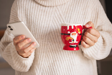 Christmas time. Closeup on woman with red santa mug in beige sweater sending text message using smartphone.